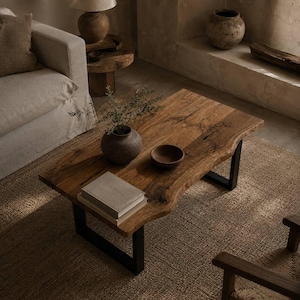 May include: A rectangular coffee table with a natural live-edge wood slab top and black metal U-shaped legs. The table surface holds a dark ceramic vase with green sprigs, a small wooden bowl, and a stack of two books with the word Kinfolk visible.