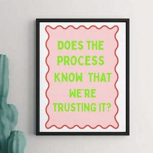Op de afbeelding: Een ingelijste kunstdruk met de tekst "DOES THE PROCESS KNOW THAT WE'RE TRUSTING IT?" in groene letters op een roze achtergrond. De roze achtergrond heeft een rode golvende rand, tegen een witte achtergrond.