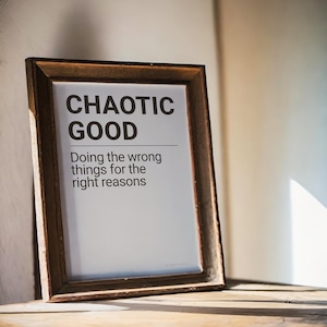 May include: A framed print with the words "CHAOTIC GOOD" in bold black letters. Below, the text reads "Doing the wrong things for the right reasons." The frame is a dark brown colour and rests on a wooden surface.
