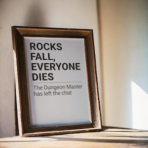 May include: A framed print with the text "ROCKS FALL, EVERYONE DIES" and "The Dungeon Master has left the chat." The frame is a dark brown color and the print is on a white background. The print is leaning against a wooden surface.