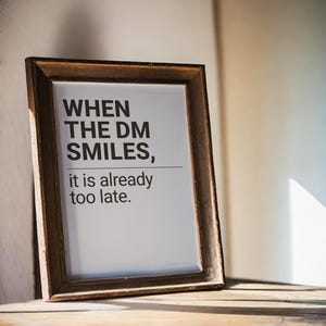Può includere: Una stampa incorniciata con il testo "WHEN THE DM SMILES, it is already too late." La cornice è di colore marrone scuro e la stampa ha uno sfondo bianco. La stampa è appoggiata a una parete su una superficie di legno.