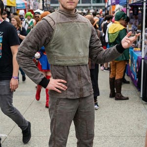 May include: A person in a brown and green costume, likely for cosplay. The outfit includes a jacket with a green vest, matching pants, and brown boots. The person is standing outside a convention center.