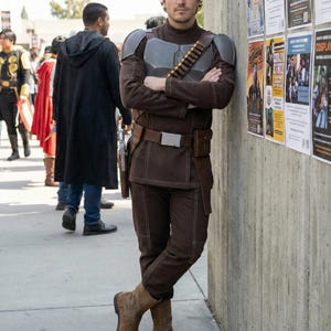 May include: A person in a brown costume with grey shoulder armour, a utility belt, and brown boots. The costume includes a bandolier and a holster. The person is leaning against a concrete wall with posters.