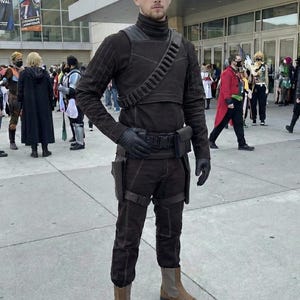 May include: A person in a brown cosplay costume, including a turtleneck, gloves, and boots. The costume features a utility belt, shoulder strap, and pouches. Other cosplayers are in the background.