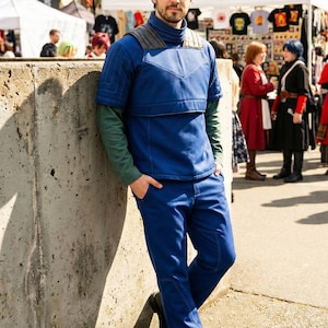 May include: A person in a blue outfit with a high collar and quilted chest piece, and matching blue trousers. The outfit has green sleeves. The person leans against a concrete wall, with a market scene in the background.