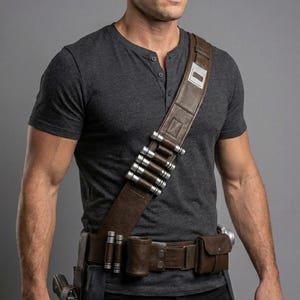 Op de afbeelding: Een bruine leren utility riem met meerdere zakjes, een holster en een beenriem. De riem wordt gedragen over een zwarte broek en een grijs Henley-shirt. De riem heeft zilverkleurige cilinders.
