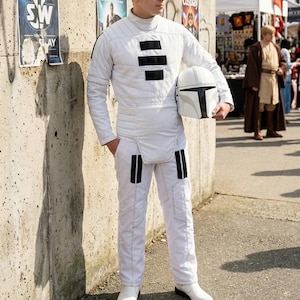 Op de afbeelding: Een persoon in een wit gewatteerd jumpsuit en laarzen, met een zwart-witte helm. De outfit heeft zwarte accenten. De persoon staat voor een betonnen muur met posters. Andere cosplayers zijn op de achtergrond.
