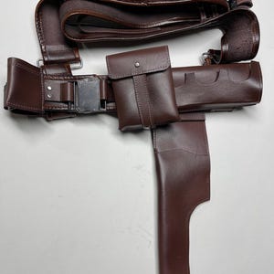 May include: A brown leather belt with multiple pouches and a holster. The belt features a silver buckle and loops for ammunition. The set includes a small pouch with a snap closure and a holster for a sidearm.