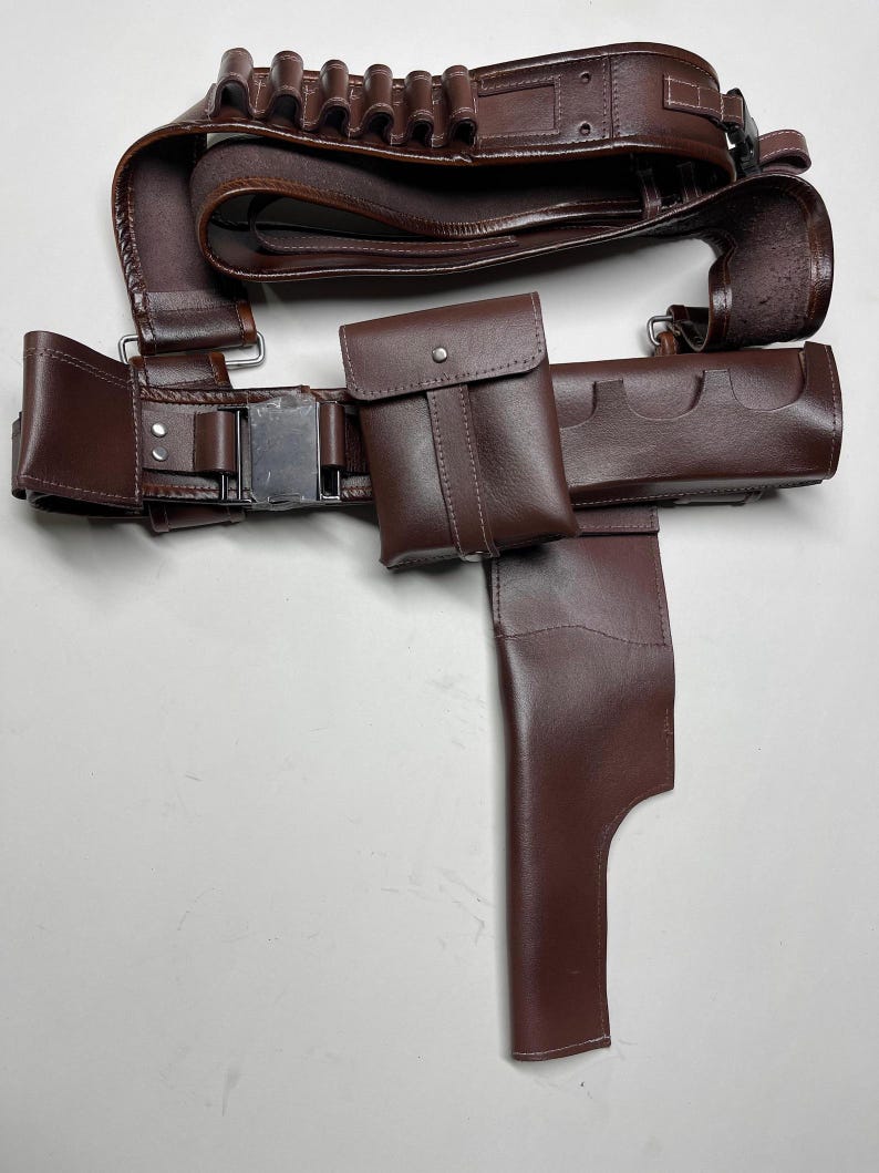 May include: A brown leather belt with multiple pouches and a holster. The belt features a silver buckle and loops for ammunition. The set includes a small pouch with a snap closure and a holster for a sidearm.
