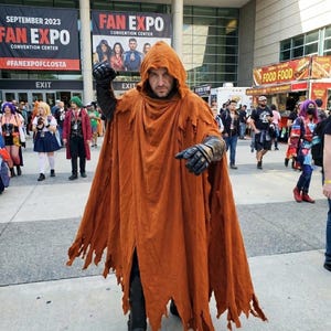 May include: A person wearing a hooded, floor-length, distressed orange cloak with frayed edges. The outfit includes black armored gloves and black boots. The cloak is draped over the shoulders and covers the torso and legs, creating a layered appearance.