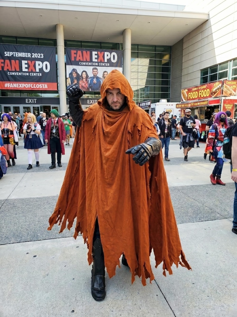 May include: A person wearing a hooded, floor-length, distressed orange cloak with frayed edges. The outfit includes black armored gloves and black boots. The cloak is draped over the shoulders and covers the torso and legs, creating a layered appearance.