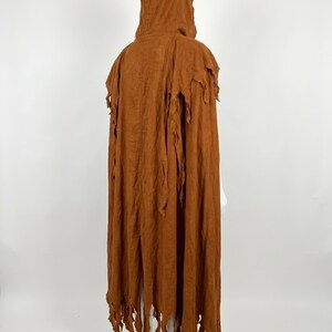 May include: A floor-length hooded cloak in a solid rust-brown color. The garment features a structured hood and a back panel with vertical seams. The hem and shoulder sections are finished with long, jagged, shredded-style fabric strips.