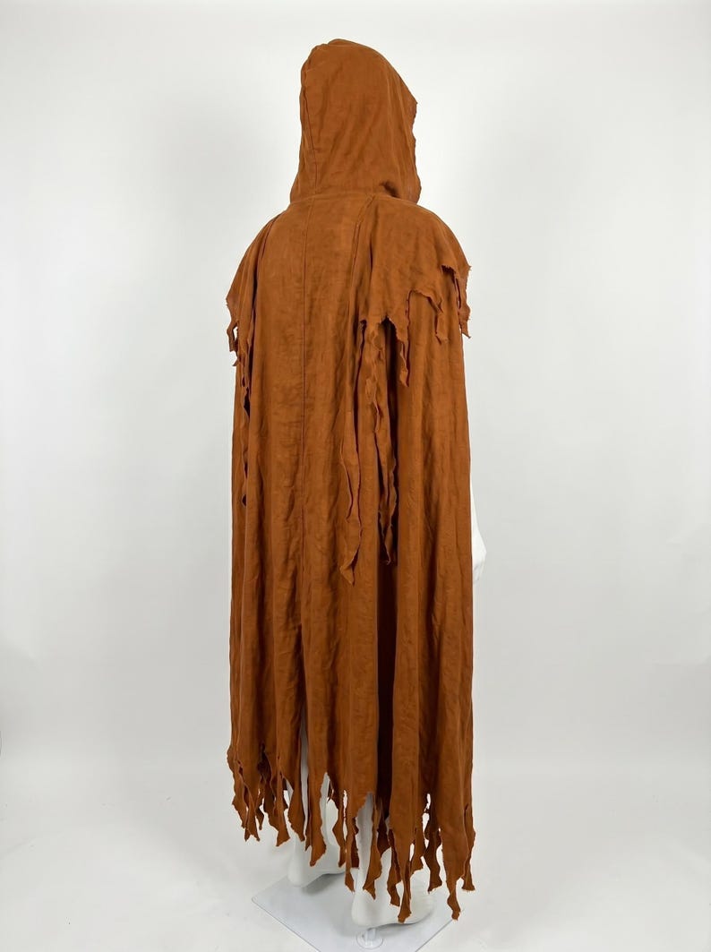 May include: A floor-length hooded cloak in a solid rust-brown color. The garment features a structured hood and a back panel with vertical seams. The hem and shoulder sections are finished with long, jagged, shredded-style fabric strips.