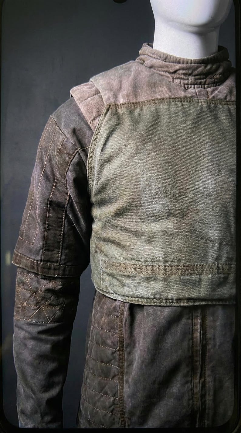 May include: A worn-looking, olive green and brown jacket with quilted detailing. The jacket has a high collar and a layered design, with a padded chest piece. The sleeves are long and feature a quilted pattern. The overall aesthetic suggests a vintage or military-inspired style.