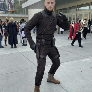 May include: A person in a brown costume with a high collar, shoulder strap, and utility belt. The costume includes gloves, pants, and boots. The person is posing in front of a building with a banner.