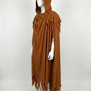 May include: A floor-length, rust-colored hooded cloak featuring a distressed, tattered hemline and shoulder edges. The garment is draped over a mannequin, showcasing a loose, flowing silhouette with a deep hood and uneven, shredded fabric details.