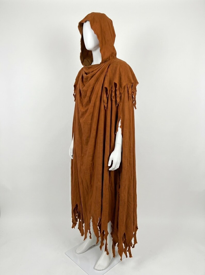 May include: A floor-length, rust-colored hooded cloak featuring a distressed, tattered hemline and shoulder edges. The garment is draped over a mannequin, showcasing a loose, flowing silhouette with a deep hood and uneven, shredded fabric details.