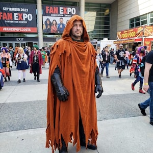 May include: A person wearing a long, distressed, burnt-orange hooded cloak with frayed edges. The costume includes black textured gauntlets on the forearms and black boots. The garment is worn over dark clothing, creating a layered appearance.