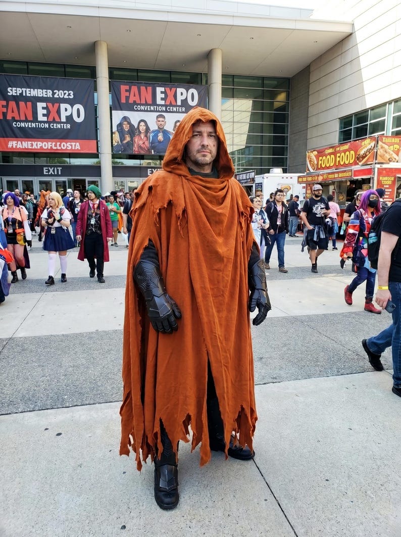 May include: A person wearing a long, distressed, burnt-orange hooded cloak with frayed edges. The costume includes black textured gauntlets on the forearms and black boots. The garment is worn over dark clothing, creating a layered appearance.