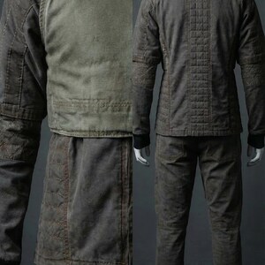 May include: A two-piece, dark gray outfit on a mannequin. The jacket has a high collar, quilted stitching, and a contrasting olive green panel. The pants are a matching dark gray with knee detailing. The outfit is designed for a rugged, utilitarian aesthetic.