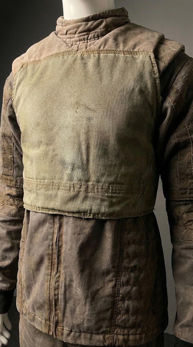 May include: A worn, brown jacket with a quilted design on the sleeves and a contrasting, lighter-colored chest panel. The jacket appears to be made of a durable, possibly canvas-like material, suggesting a workwear or utilitarian aesthetic.