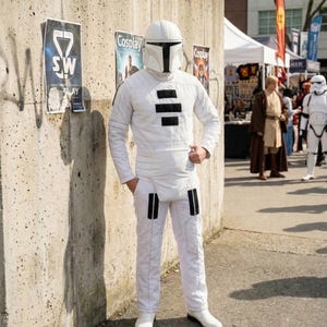 Op de afbeelding: Een persoon in een wit Mandalorian cosplay kostuum staat bij een betonnen muur met posters. Het kostuum omvat een helm, jas met lange mouwen en broek, allemaal in wit met zwarte accenten. De persoon draagt witte laarzen.
