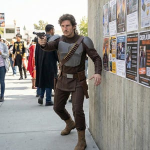 May include: A person in a brown and gray costume, holding a gun and aiming. The costume includes a chest plate, belt, and boots. The background shows a concrete wall with posters and other people in costumes.