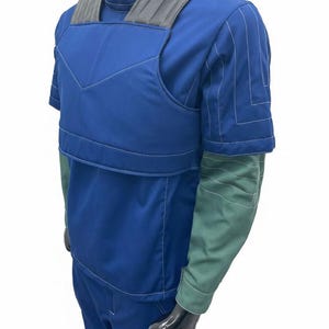 May include: Blue and green short-sleeve jumpsuit with gray shoulder padding. The jumpsuit has white stitching details and a high collar. The sleeves are green, and the body is blue. The jumpsuit is displayed on a black mannequin.