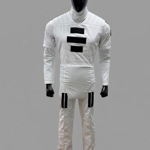 Op de afbeelding: Een witte jumpsuit met zwarte accenten, gepresenteerd op een mannequin. De outfit heeft een hoge kraag, lange mouwen en zwarte rechthoekige details op de borst en benen. Het pak is ontworpen met gewatteerde stiksels.