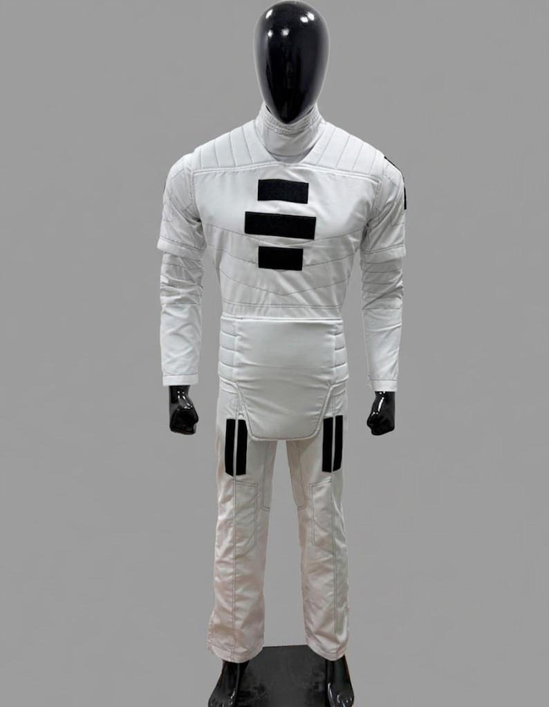 Op de afbeelding: Een witte jumpsuit met zwarte accenten, gepresenteerd op een mannequin. De outfit heeft een hoge kraag, lange mouwen en zwarte rechthoekige details op de borst en benen. Het pak is ontworpen met gewatteerde stiksels.