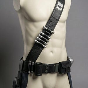 Op de afbeelding: Zwarte leren utility riem en schouderriem met zilverkleurige accenten. De riem heeft meerdere zakjes, een holster en een messenschede. De schouderriem heeft een rechthoekig zilverkleurig detail en een rij zilverkleurige cilinders.