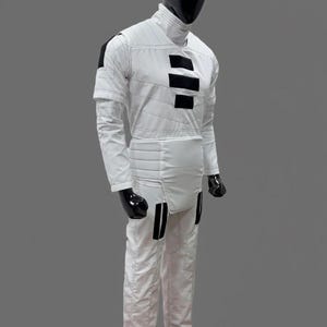 Op de afbeelding: Een witte jumpsuit met zwarte accenten, getoond op een mannequin. De outfit heeft een hoge kraag, horizontale zwarte strepen op de borst en zwarte details op de schouders en benen. De mannequin heeft een zwarte kop en handschoenen.