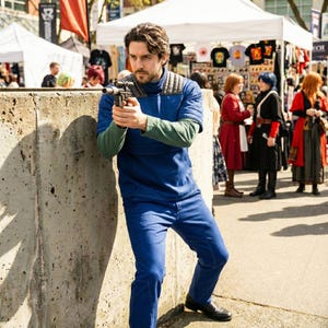 May include: A person in a blue jumpsuit and green undershirt holds a prop gun, leaning against a concrete wall. The scene is set outdoors, with a crowd and vendor tents in the background. The person is wearing black shoes.