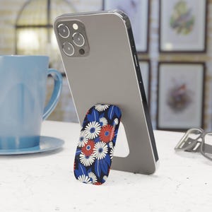Può includere: Uno smartphone sostenuto da un supporto per telefono con motivo floreale. L'impugnatura presenta uno sfondo blu navy con fiori e foglie bianche, rosse e blu a forma di margherita. Una tazza blu è sullo sfondo.