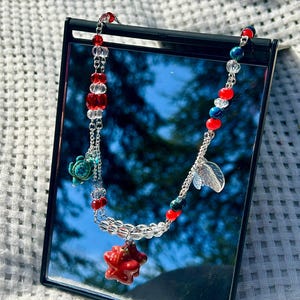 May include: A necklace featuring red, white, and blue beads, a red star pendant, and a small turquoise turtle charm. The necklace is displayed on a black mirror, reflecting a blurred outdoor scene.