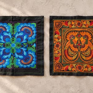May include: Two square black fabric panels featuring intricate embroidery. The left panel displays a symmetrical blue and green floral pattern. The right panel features an orange and yellow bird motif surrounded by decorative floral borders.