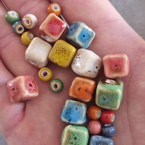 Op de afbeelding: Een verzameling kleine keramische kralen in diverse vormen en kleuren. Het assortiment bevat vierkante en ronde kralen met gespikkelde, geglazuurde afwerkingen in tinten blauw, groen, geel, oranje, roze en wit, elk voorzien van een centraal gat.
