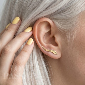 May include: A gold-colored ear climber earring in the shape of a feather is worn on an ear. The earring curves along the ear's edge. The person's nails are painted yellow.