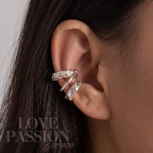 May include: A set of three silver-toned ear cuffs worn on the cartilage. The top cuff features a textured, cratered surface, the middle cuff is a smooth, polished band, and the bottom cuff has an irregular, hammered finish with a reflective sheen.