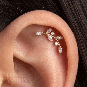 May include: A gold-toned ear cartilage piercing jewellery piece shaped like a curved branch with multiple marquise-cut and round-cut clear stones arranged along the stem. The jewellery is positioned along the upper curve of the ear cartilage.