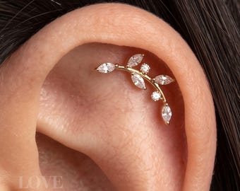 Può includere: Un gioiello per piercing alla cartilagine dell'orecchio color oro a forma di ramo curvo con diverse pietre trasparenti a taglio marquise e rotondo disposte lungo il gambo. Il gioiello è posizionato lungo la curva superiore della cartilagine.