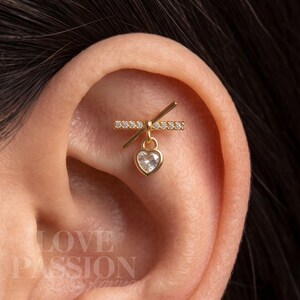 May include: A gold-toned ear piercing jewelry piece featuring a horizontal bar encrusted with small clear stones, crossed by a thin diagonal gold bar, with a dangling heart-shaped charm containing a single clear stone in the center.