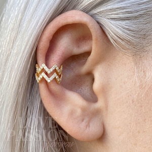 May include: Gold-toned ear cuff with a zigzag design, encrusted with small, sparkling crystals. The cuff wraps around the ear, creating a layered effect. The jewelry piece is shown on an ear with gray hair in the background.