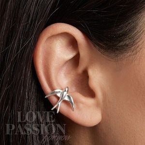 Peut inclure: Une boucle d'oreille en argent en forme d'oiseau en plein vol, présentant des surfaces métalliques lisses et polies ainsi que des contours d'ailes détaillés. L'oiseau est placé sur le bord extérieur du cartilage de l'oreille.