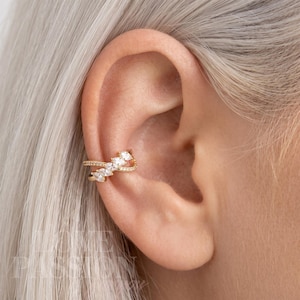May include: Gold-toned ear cuff featuring a delicate design. The cuff wraps around the ear with a series of sparkling, clear gemstones. The design includes a row of smaller stones and larger, marquise-shaped gems, creating a sophisticated look.