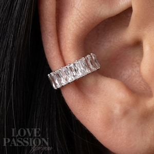 May include: A silver-toned ear cuff featuring a row of clear, rectangular baguette-cut stones set in a metallic band. The jewellery piece is positioned along the outer edge of the ear cartilage, showcasing a series of faceted, sparkling crystals.