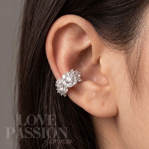 May include: A silver-toned ear cuff featuring a cluster of multiple round, clear, faceted stones arranged in a dense, sparkling formation. The jewellery piece wraps around the cartilage of the ear, showcasing a bright, reflective metallic finish.