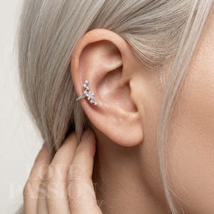 Puede incluir: Un ear cuff plateado con un diseño floral con múltiples piedras preciosas transparentes. El brazalete se curva a lo largo de la oreja, con las piedras preciosas agrupadas para crear un efecto brillante. La joya se muestra en una persona con cabello claro.