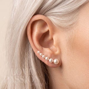 Pode incluir: Um brinco ear climber com uma fileira de seis pérolas esféricas brancas de tamanhos graduados, seguindo a curva natural do lóbulo da orelha. As pérolas estão dispostas da menor para a maior, criando um design linear ao longo da orelha.