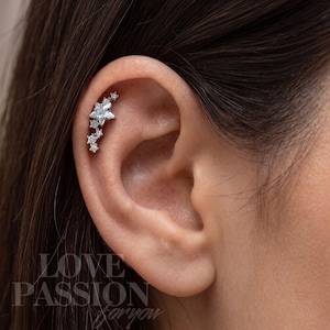 May include: A silver-toned ear cartilage piercing featuring a curved cluster of clear, faceted stones arranged in a star-like pattern. The jewelry follows the natural curve of the upper ear, showcasing multiple small round stones and one larger central star-shaped stone.